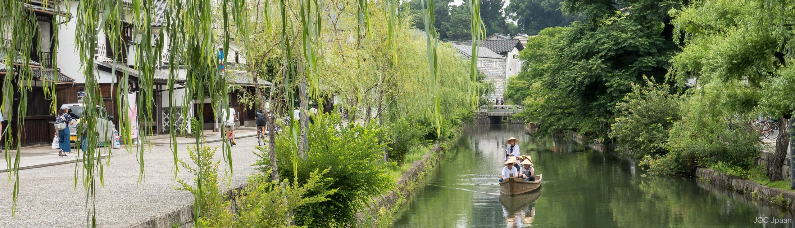 A tranquil Kurashiki canal lined with greenery and willow trees, where people in traditional hats ride a wooden boat and pedestrians stroll beside historic buildings along the water.