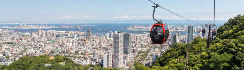 Red cable cars travel above a lush green hillside in Kobe, overlooking a sprawling cityscape and harbor under a clear blue sky.