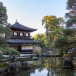 Ginkakuji Temple (銀閣寺) – Kyoto’s Silver Pavilion A traditional Japanese pavilion, reminiscent of Kyoto’s Ginkakuji Temple (Silver Pavilion), stands beside a serene pond, surrounded by lush trees and rocks in a peaceful garden under a clear blue sky.