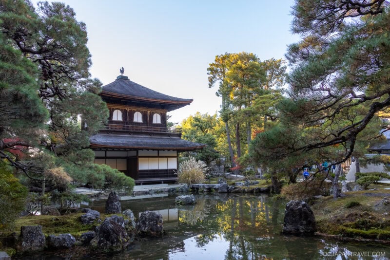A traditional Japanese pavilion, reminiscent of Kyoto&rsquo;s Ginkakuji Temple (Silver Pavilion), stands beside a serene pond, surrounded by lush trees and rocks in a peaceful garden under a clear blue sky.