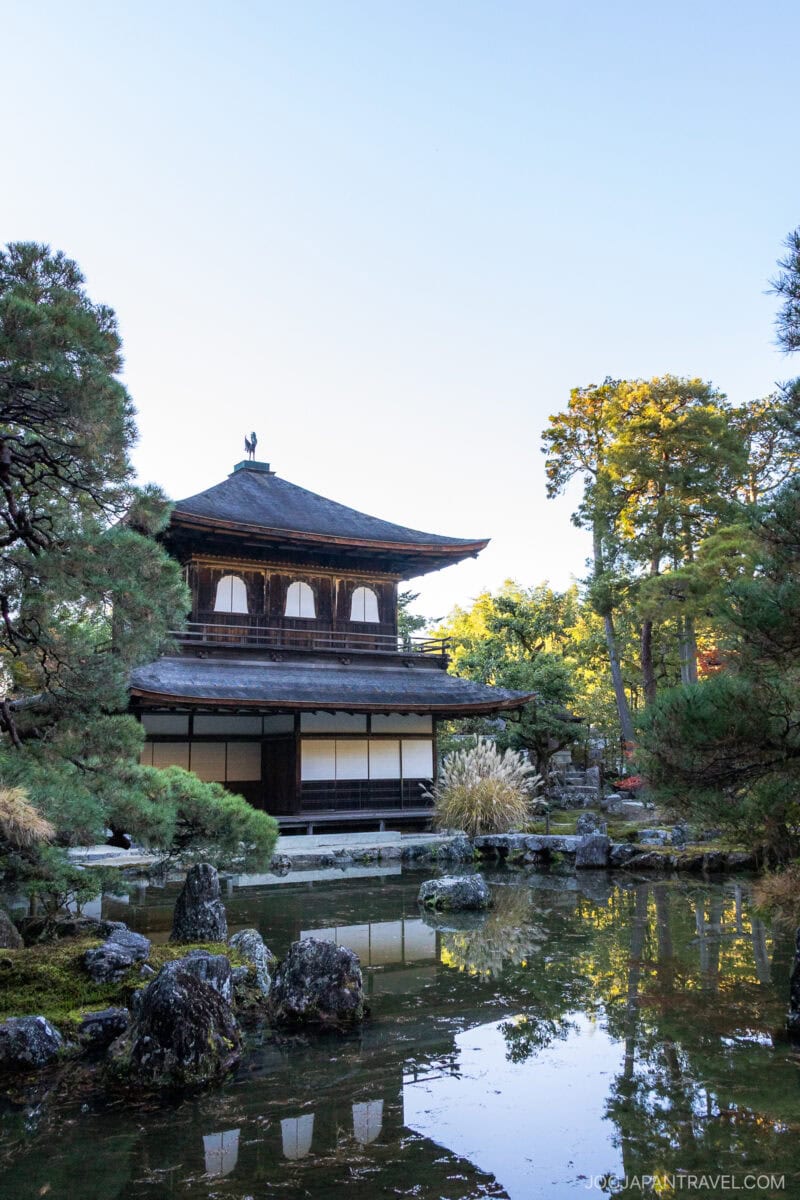The Ginkakuji Temple, also known as the Silver Pavilion, stands by a tranquil pond surrounded by trees and rocks under a clear blue Kyoto sky.