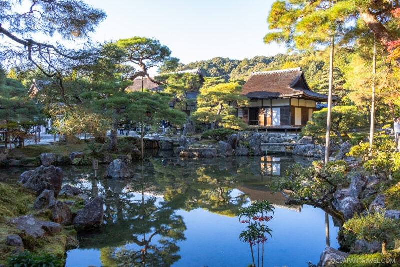 A serene Japanese garden at Kyoto’s Ginkakuji Temple features a pond reflecting green trees and traditional wooden buildings under a clear blue sky, surrounded by lush greenery and distant hills.