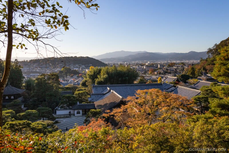 View from a hillside overlooking Kyoto, Japan, with traditional temple rooftops like Ginkakuji Temple (Silver Pavilion), lush green and autumn foliage in the foreground, and mountains in the distance under a clear sky.