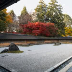 Ryoan-ji (竜安寺): Kyoto’s Mysterious Zen Rock Garden A traditional Japanese Zen rock garden at Ryoan-ji features raked gravel, several large rocks, and a backdrop of trees with autumn foliage in green, yellow, and red hues. The serene garden is enclosed by classic wooden walls.