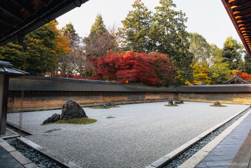 A traditional Japanese Zen rock garden at Ryoan-ji features raked gravel, several large rocks, and a backdrop of trees with autumn foliage in green, yellow, and red hues. The serene garden is enclosed by classic wooden walls.