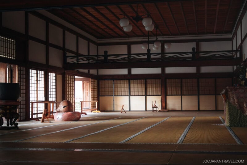 A spacious, traditional Japanese tatami room inspired by Tenryu-ji Temple, featuring wooden beams, shoji screens, paper lanterns, and minimal furnishings. A large drum and decorative mask rest on the floor as natural light filters through the windows.