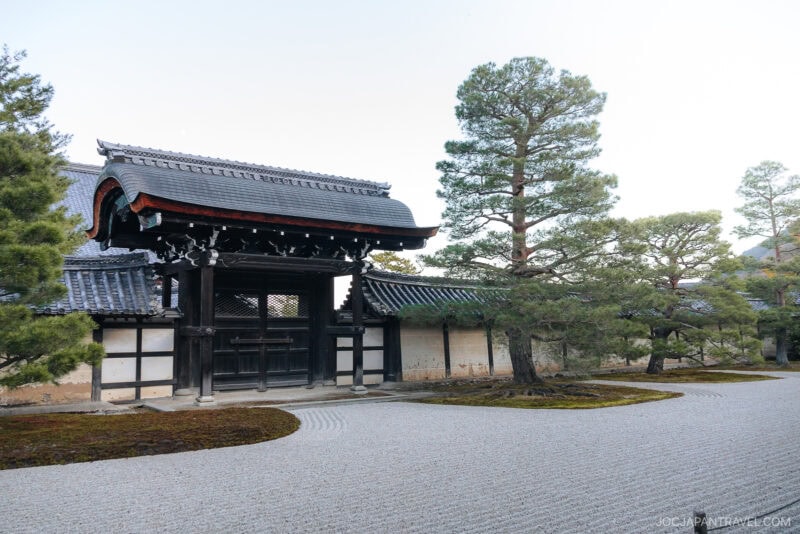 A traditional Japanese wooden gate at Tenryu-ji Temple stands before a stone wall, surrounded by neatly trimmed pine trees and a raked gravel garden. The scene is tranquil and well-maintained.
