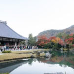Tenryu-ji Temple (天龍寺): Kyoto’s Zen Sanctuary and Historic Gardens A traditional Japanese building at Tenryu-ji Temple stands beside a reflective pond, with people walking nearby and colorful autumn trees and forested hills in the background.