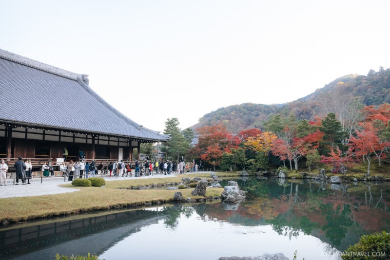 A traditional Japanese building at Tenryu-ji Temple stands beside a reflective pond, with people walking nearby and colorful autumn trees and forested hills in the background.