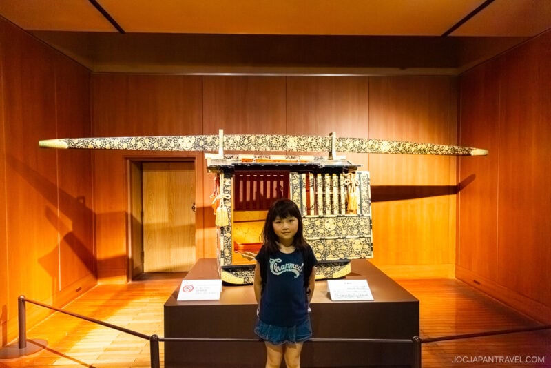 A young girl stands in front of a traditional Japanese palanquin display at a museum near Nagoya Castle, with warm wooden walls and an informational sign nearby.