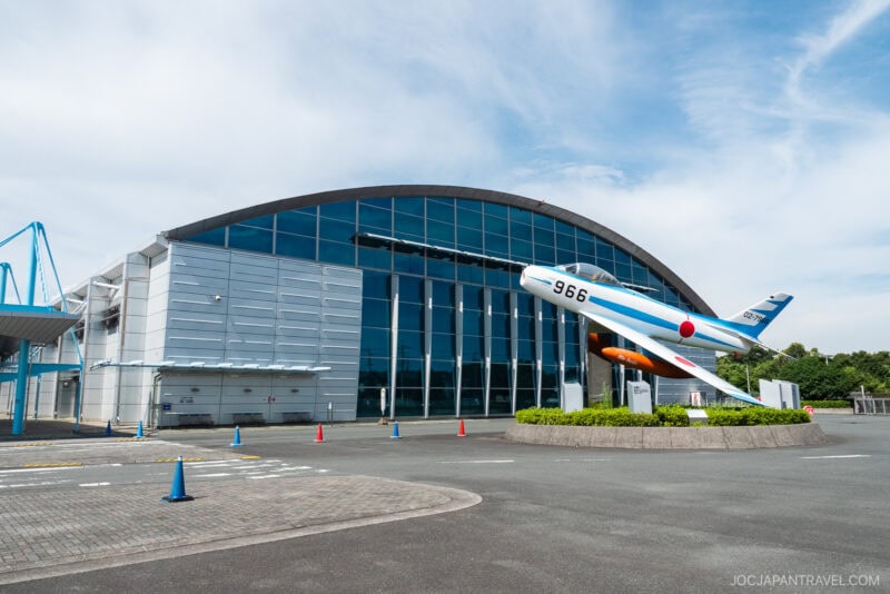 A modern glass-fronted aviation museum, Airpark Hamamatsu features a large fighter jet and missile on pedestals outside; blue sky and a few traffic cones are visible in the foreground.