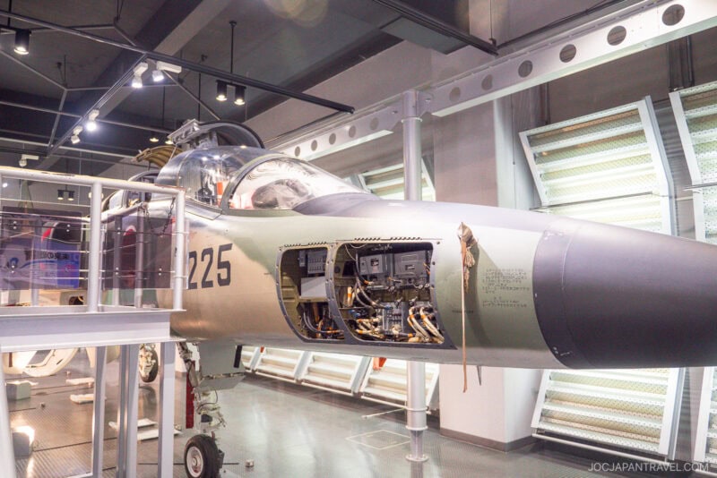A military jet is displayed at Airpark Hamamatsu, with panels open to reveal internal electronics and wiring. The aircraft, marked with the number 225, is surrounded by railings and modern lighting.
