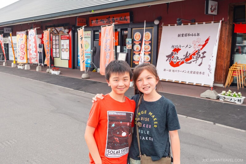 Two smiling children stand close together in front of a Japanese restaurant at Asahikawa Ramen Village, with bright banners, menus, and a row of flowerpots along the sidewalk. The boy wears a red T-shirt, and the girl wears a dark T-shirt.