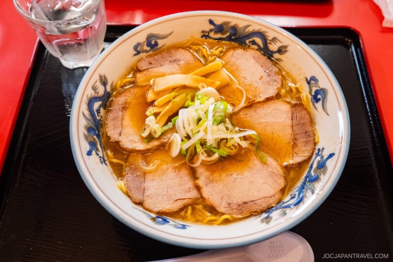 A bowl of ramen from Asahikawa Ramen Village, topped with pork slices, bamboo shoots, and chopped green onions, sits on a black tray next to a glass of water and a white spoon.