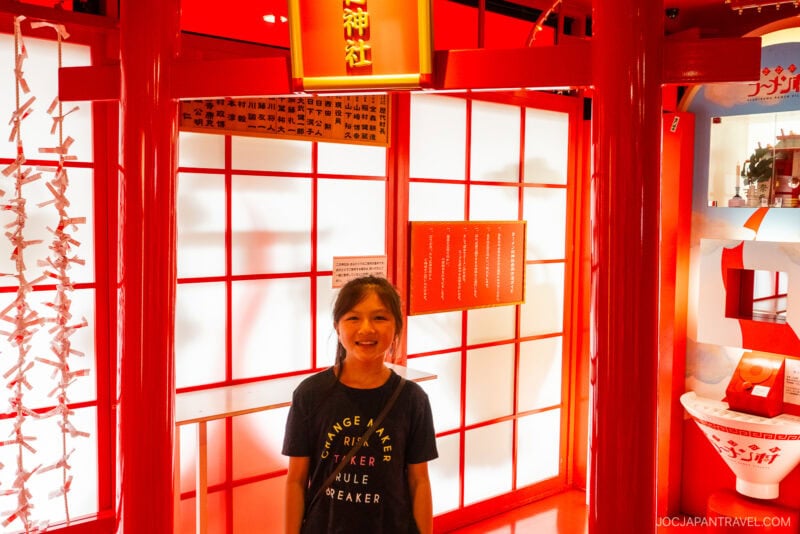 A smiling girl stands in front of a bright red and white display with Japanese writing, featuring traditional decor and folded paper strips, likely at Asahikawa Ramen Village or another cultural attraction.
