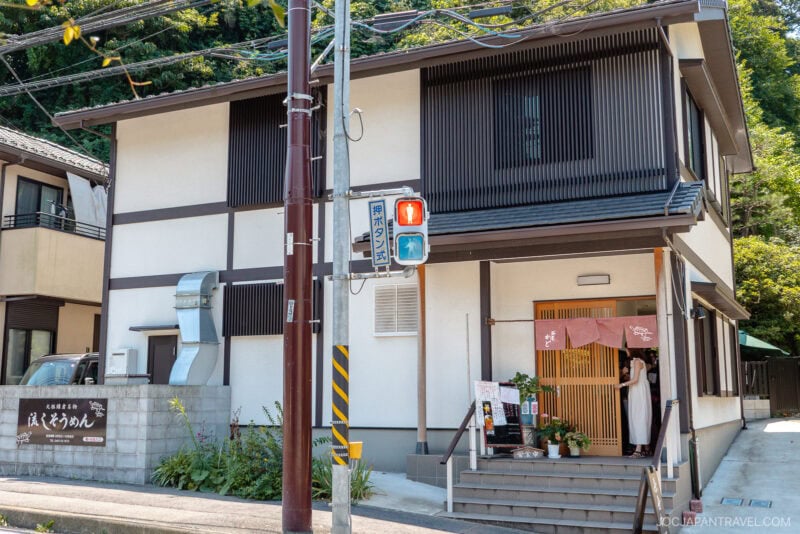 A modern two-story Japanese building with vertical wooden slats, a small Chayakado restaurant entrance draped in noren curtain, and potted plants. Two people stand at the entrance near a red traffic light.