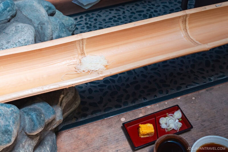 A few strands of somen noodles rest in a bamboo chute for nagashi somen at Chayakado, with dipping sauce, sliced green onions, and tamagoyaki in small dishes nearby on a wooden table.