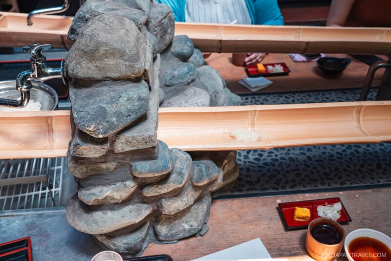 At Chayakado, a bamboo water slide for nagashi somen winds over rocks, with plates of dipping sauces and condiments on the table. People sit around, ready to catch noodles as they flow down the bamboo chute.