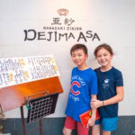 Dejima Asa (出島亜紗) – Sampling the Regional Flavors of Nagasaki Two children stand smiling in front of a restaurant sign that reads Dejima Asa Nagasaki Dining. A menu with Japanese writing is displayed on a stand next to them, inviting guests to explore the flavors of Dejima Asa.