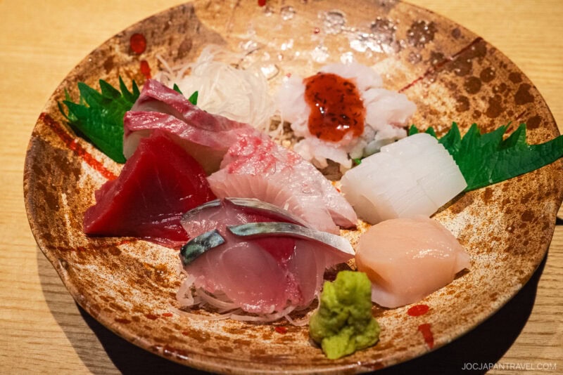 A ceramic plate with assorted sashimi—including Dejima Asa tuna, mackerel, squid, scallop, and white fish—garnished with shiso leaves, daikon radish, wasabi, and a dollop of red sauce on a wooden table.