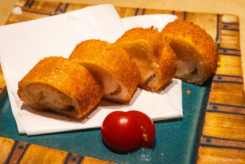 Four pieces of golden-brown, breaded and fried Dejima Asa are arranged on a napkin, next to a halved cherry tomato, on a decorative blue and tan plate.