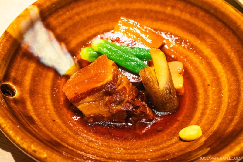 A brown ceramic bowl from Dejima Asa holds braised pork belly in a rich sauce, garnished with green beans, carrots, bamboo shoots, and a single peanut. The dish is brightly lit and attractively presented.