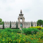 Exploring Arita (有田町) – The Birthplace of Japanese Porcelain An ornate, historic palace with a large crown-shaped spire stands behind lush gardens filled with colorful flowers and green shrubs, while delicate arita porcelain accents adorn the scene under a cloudy sky.