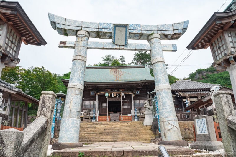A weathered stone torii gate stands before the entrance to a traditional Japanese shrine in Arita, with stone steps leading up to the main building surrounded by wooden structures and lush greenery.