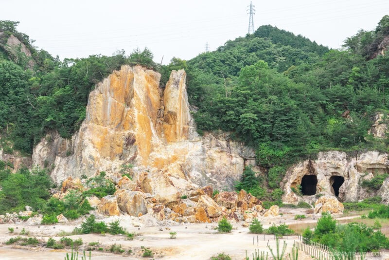 A rocky cliff with yellowish-orange hues stands beside two large cave entrances, surrounded by scattered boulders, arita greenery, and a forested hillside in the background under a cloudy sky.
