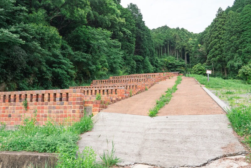 A sloped path with red brick walls on each side leads into a lush, green forest near Arita. The area is surrounded by dense trees and vegetation, and a small sign is visible on the right side of the path.