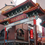 Nagasaki Shinchi Chinatown (長崎新地中華街) – Japan’s Oldest Chinatown A traditional Chinese-style entrance gate with ornate details and red columns stands in an urban area, marking the entrance to Nagasaki Shinchi Chinatown. Surrounding buildings display both Japanese and Chinese signage.