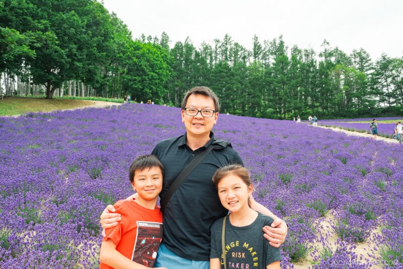 A man stands smiling between two children in front of a large lavender field at Farm Tomita, with tall green trees in the background under a bright, clear sky.