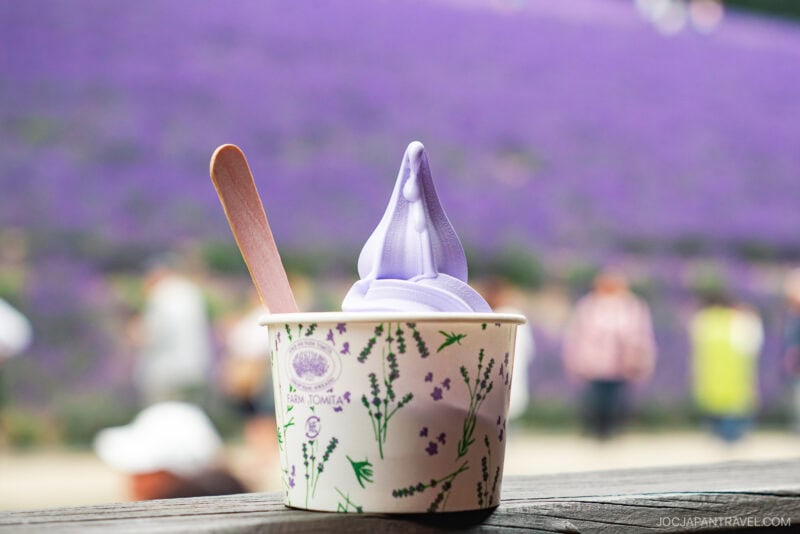 A cup of lavender-colored soft serve ice cream from Farm Tomita with a wooden spoon sits on a table, with a blurred lavender field and people in the background. The cup features a delicate floral lavender design.