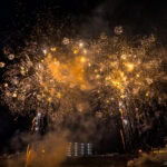 Fireworks in Izunokuni (伊豆の国市) – Summer Festival Magic Bright golden fireworks burst in the night sky above silhouetted buildings at the Izunokuni Fireworks Festival, with smoke drifting across the scene. The lights illuminate the dark, festive atmosphere below.