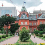 Former Hokkaido Government Office (北海道庁旧本庁舎) – Sapporo’s Red Brick Landmark The Former Hokkaido Government Office is a historic red-brick building with a dome, nestled among gardens, trees, and pathways where people stroll; modern buildings and cranes rise in the background.