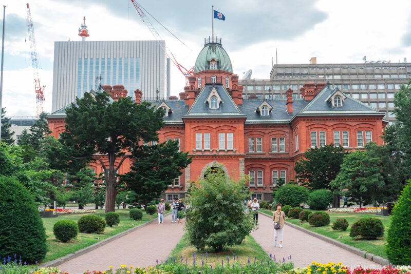 The Former Hokkaido Government Office is a historic red-brick building with a dome, nestled among gardens, trees, and pathways where people stroll; modern buildings and cranes rise in the background.