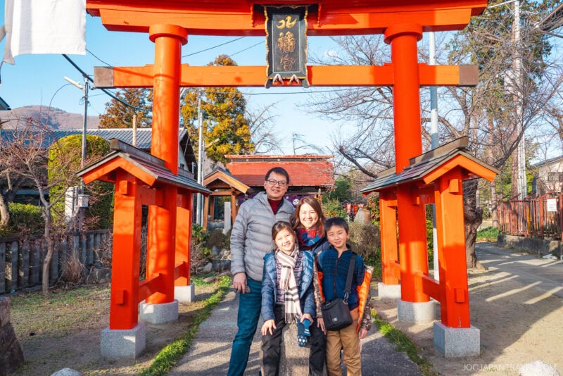 A smiling family of four poses in front of a bright red torii gate at a Fuefuki shrine on a sunny day, with trees and traditional buildings in the background.