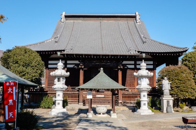 A traditional Japanese temple in Fuefuki with a tiled roof, ornate wooden facade, two white stone lanterns, and a small pavilion in front, surrounded by trees and a red banner on the left.