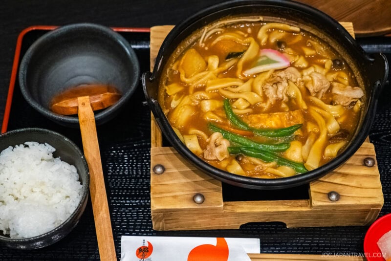 A bowl of steaming hot udon noodle soup with vegetables and meat in a pot, inspired by Fuefuki flavors, served with white rice, a wooden spoon, and chopsticks on a tray.