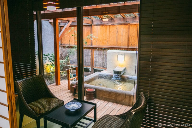 A cozy room in Fuefuki with two wicker chairs and a small table overlooks a private outdoor Japanese onsen bath, surrounded by wooden walls and greenery, bathed in warm ambient lighting.