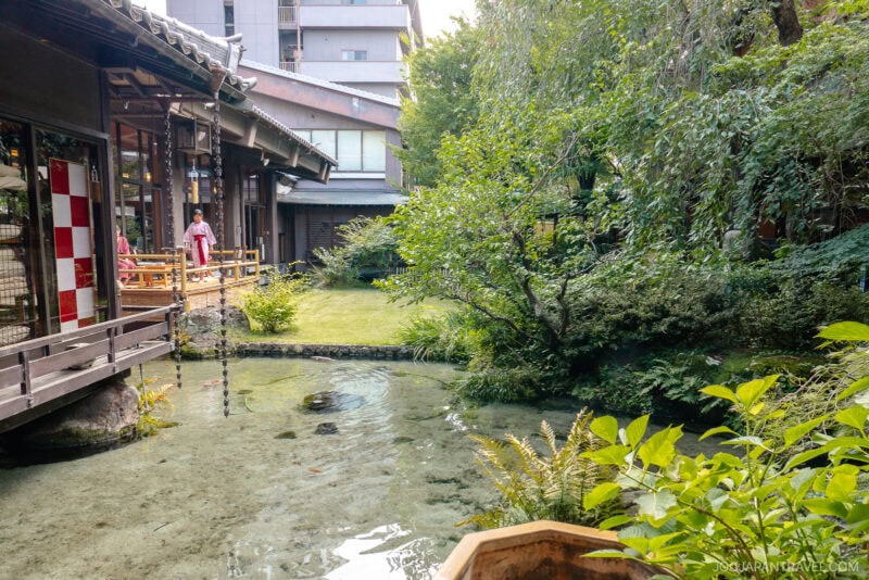 A traditional Fuefuki Japanese building stands beside a clear pond and lush garden; a person in a kimono graces the wooden terrace, with greenery, a lawn, and another building visible in the background.