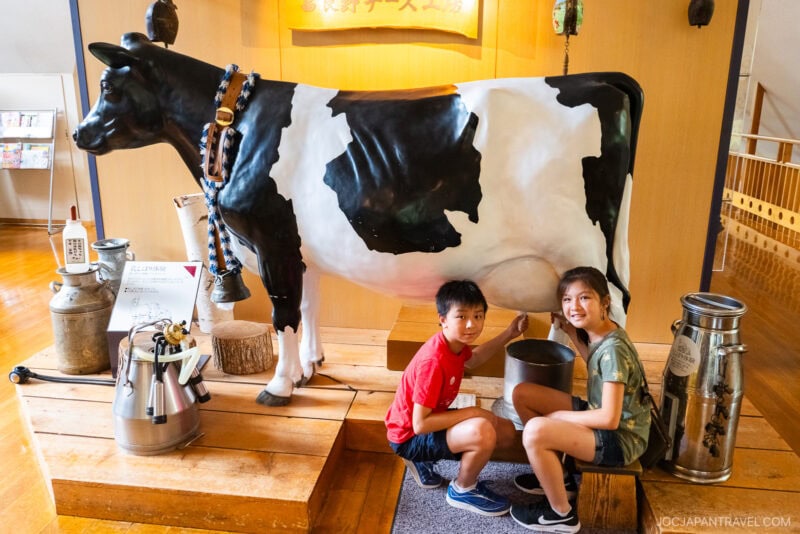 Two smiling children kneel beside a life-sized black and white cow statue indoors at the Furano Cheese Factory, pretending to milk it. Various milk cans and buckets are displayed nearby on wooden platforms.