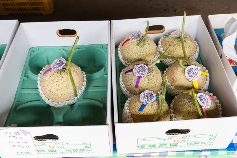 Boxes of Furano Melon cantaloupes with stems attached, each fruit wrapped in a white protective net and labeled with colorful tags, displayed for sale at a market.
