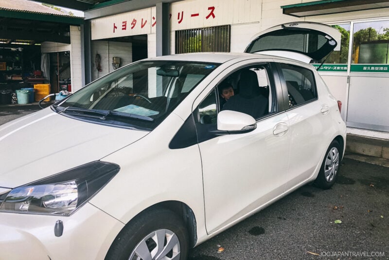 A white car with an open hatchback is parked outside a building with Japanese signs in Yakushima. A person sits in the driver’s seat, partially visible through the window. The car appears ready for travel or loading.