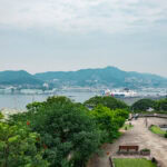 Glover Garden – Western Homes and Festival Culture in Nagasaki A scenic view of a harbor with a large cruise ship docked, surrounded by green hills, city buildings, and Glover Garden. People walk along a paved park pathway in the foreground, with benches and lush trees.