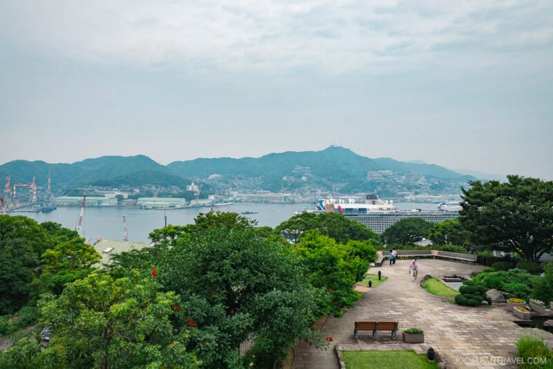 A scenic view of a harbor with a large cruise ship docked, surrounded by green hills, city buildings, and Glover Garden. People walk along a paved park pathway in the foreground, with benches and lush trees.