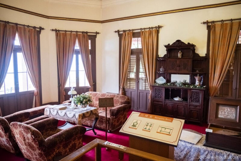 A historic Glover Garden reception room with plush armchairs, a sofa, lace-covered tables, a wooden cabinet displaying ceramics, and gold curtains on tall windows. Informational plaques and a red carpet add to the vintage ambiance.
