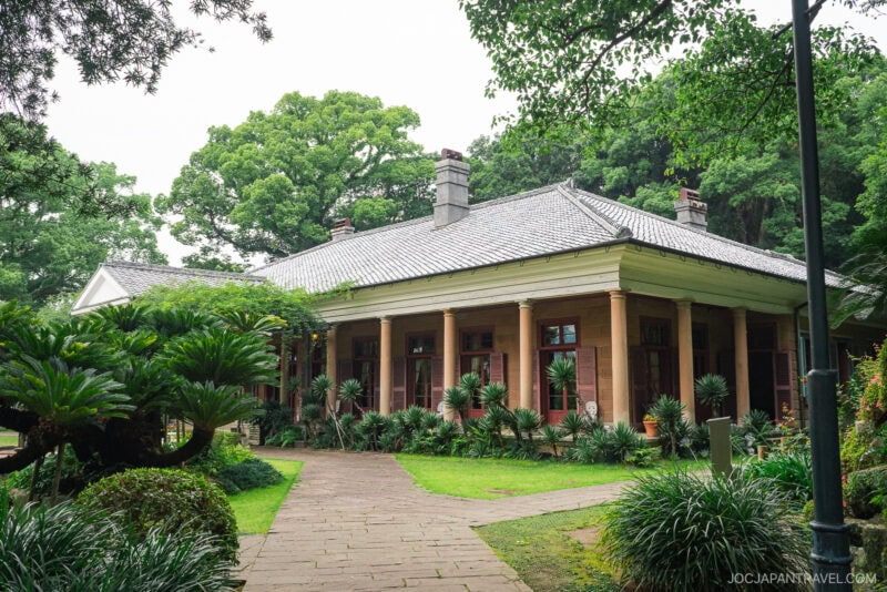 A historic Western-style building with tall columns and a tiled roof stands amidst lush greenery and tropical plants in Glover Garden, all beneath a cloudy sky.