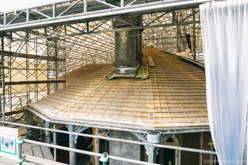 A dome-shaped roof in Glover Garden under renovation, surrounded by scaffolding and partially covered by a transparent tarp, with a central chimney and exposed wooden shingles.