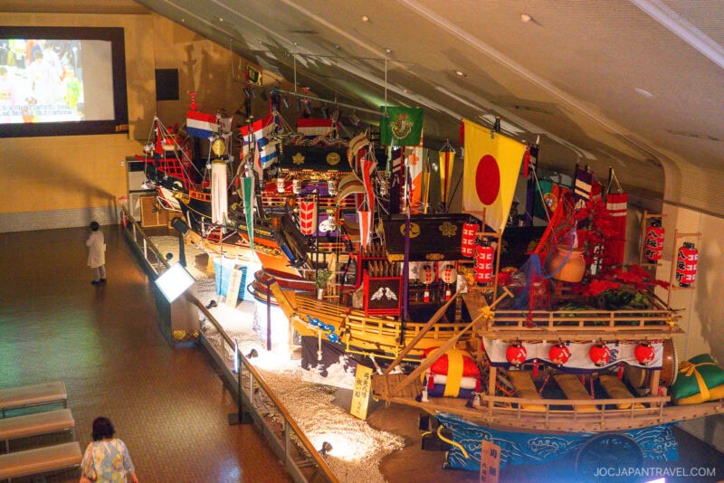A museum exhibit features a large, colorful traditional Japanese ship adorned with flags, lanterns, and decorations—reminiscent of vessels seen at Glover Garden. Visitors walk nearby, and a video screen glows on the wall in the background.